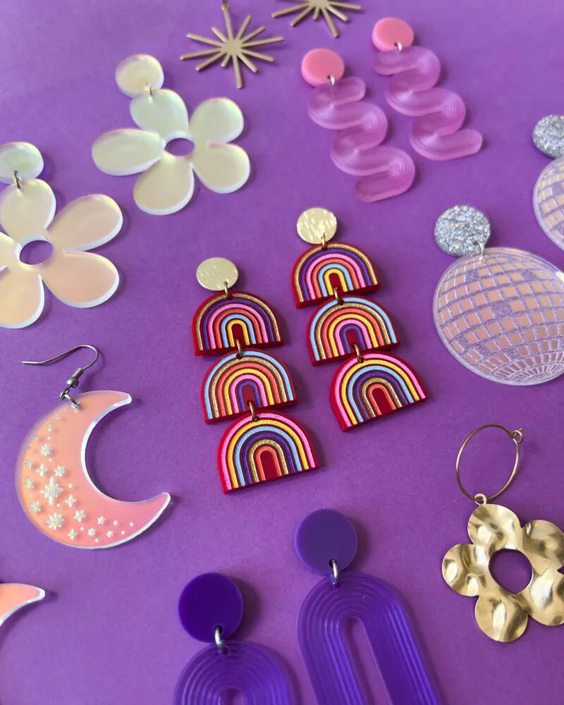 Colorful handmade earrings spread on a purple surface, including rainbow, floral, moon, and disco ball styles