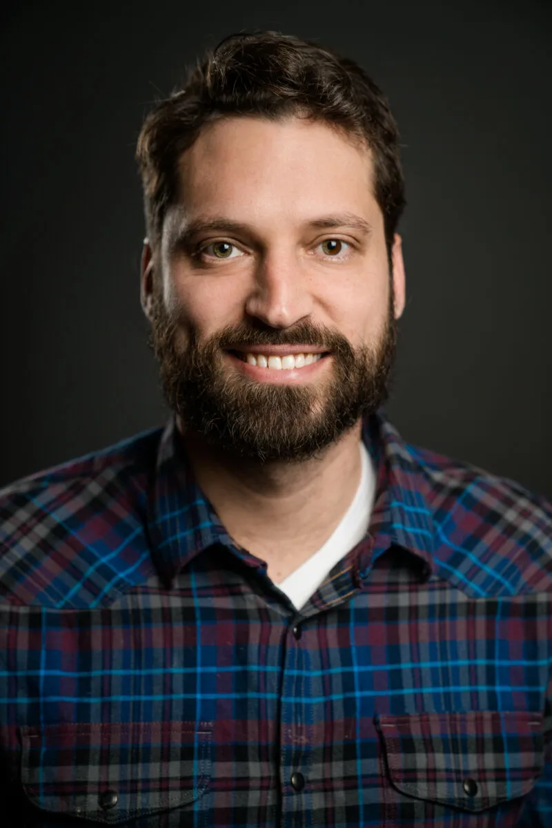 Smiling bearded man in a blue plaid flannel shirt against a dark studio background