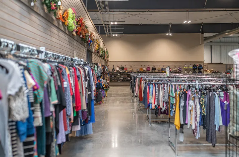 Rows of colorful clothing racks fill a spacious thrift store interior with handbags displayed along the back wall