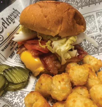 Bacon cheeseburger with lettuce and tomato beside tater tots and pickles on newsprint