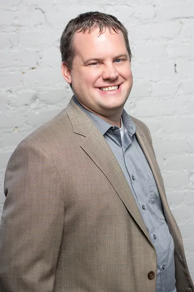 Smiling man in a tan blazer and gray shirt against a white brick wall