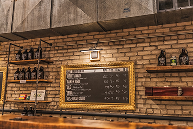 Craft brewery tap room with brick wall, gold-framed chalkboard beer menu, and bottle-lined shelves