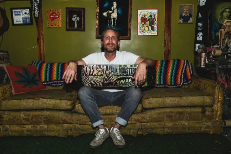 Man in white tee holding a decorated skateboard on a worn couch surrounded by eclectic wall art