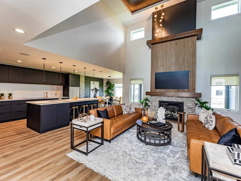 Open-concept living room with tan leather sofas, stone fireplace, and dark-cabinet kitchen beyond