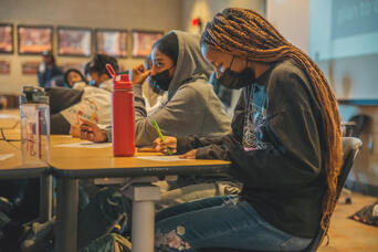 Students wearing masks write at desks in a dimly lit classroom