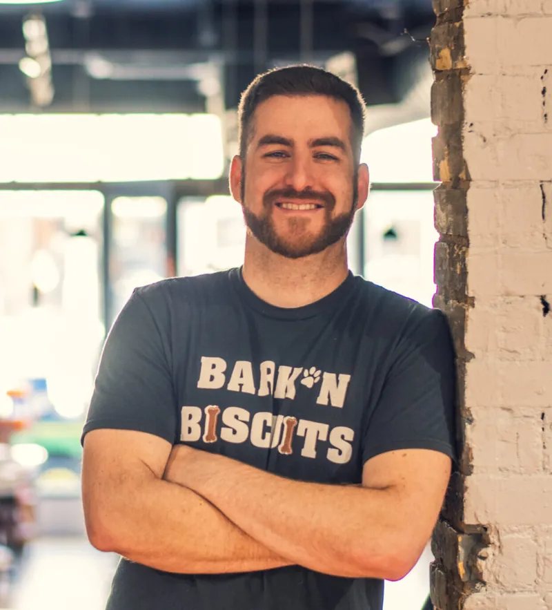 Smiling bearded man with arms crossed wearing a Bark'n Biscuits T-shirt, leaning against a white brick column