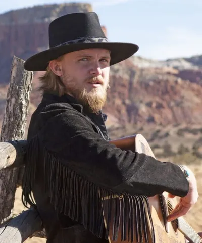 Blond bearded man in black wide-brim hat and fringed jacket leaning on a fence with red rock mesas behind him