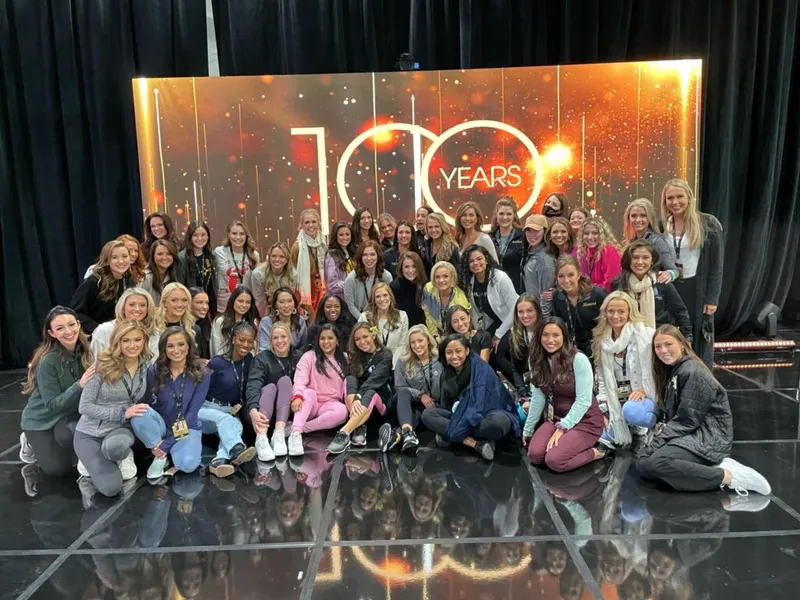 Large group of young women posing in front of a glittering '100 Years' backdrop on a stage