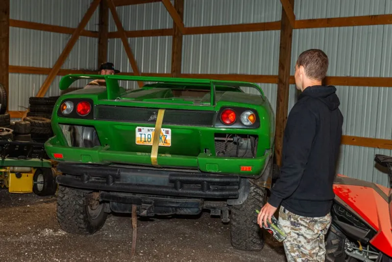 Two men inspect the rear of a bright green sports car on oversized tires inside a metal barn