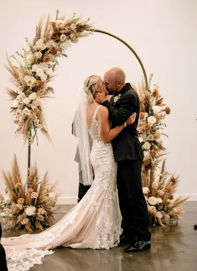 Bride and groom sharing first kiss beneath a circular floral arch with pampas grass and blush roses