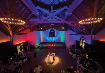 Couple sharing first dance in a rustic barn venue with colorful LED stage lighting and exposed wood-beam ceiling