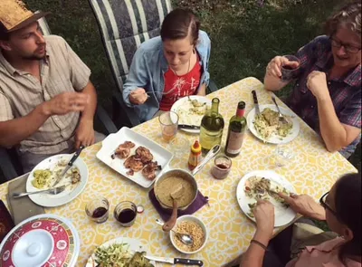 Four people sharing an outdoor meal around a yellow floral tablecloth with wine, grilled meat, and salads