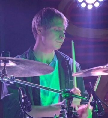 Young male drummer mid-performance under green and purple stage lights