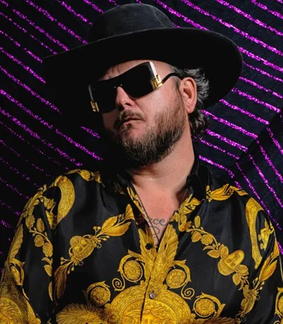Bearded man in oversized gold-frame sunglasses, black wide-brim hat, and black-and-gold baroque shirt