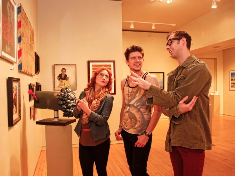 Three young adults smiling and chatting beside a sculpture in a gallery lined with framed artwork