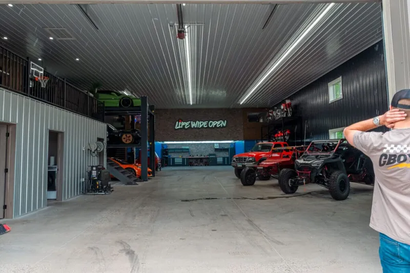 Spacious garage interior with off-road trucks, a car lift, and a neon 'Life Wide Open' sign