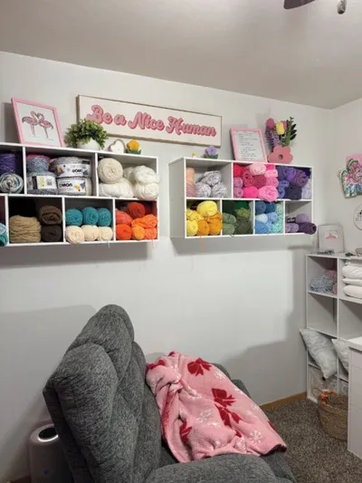 Colorful yarn balls arranged on white wall shelves above a gray sofa, with a 'Be a Nice Human' sign