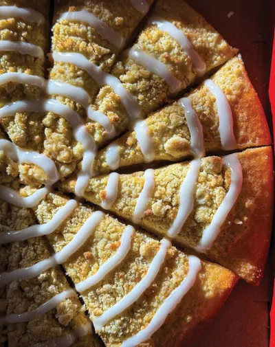 Round crumb-topped cake drizzled with white icing, cut into wedges