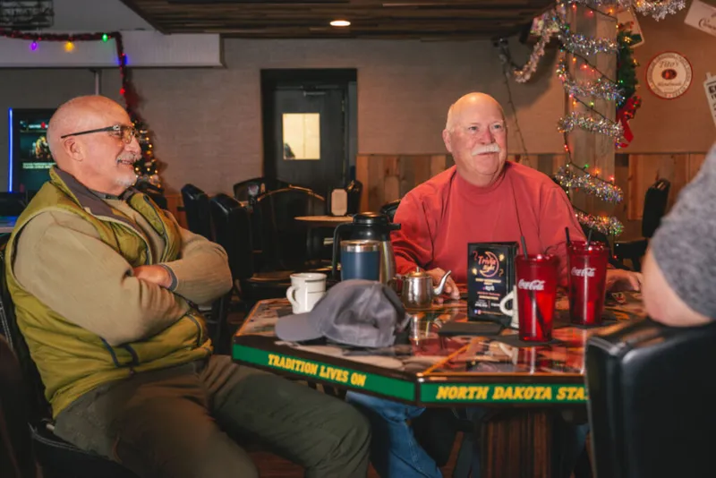 Two older men chatting at a bar table with a North Dakota State-themed edge and red Coca-Cola cups