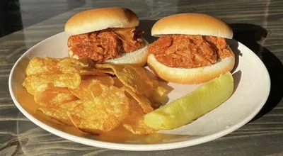 Two pulled pork sliders on a white plate with potato chips and a pickle spear