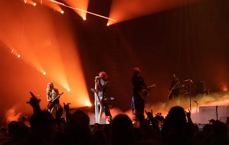 Rock band performing on a smoke-filled stage bathed in orange and red stage lighting before a crowd