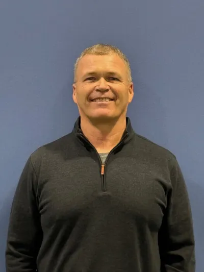 Smiling middle-aged man with short blond hair wearing a dark quarter-zip pullover against a blue wall
