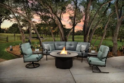 Patio seating set with curved sofa and armchairs around a round gas fire pit at dusk