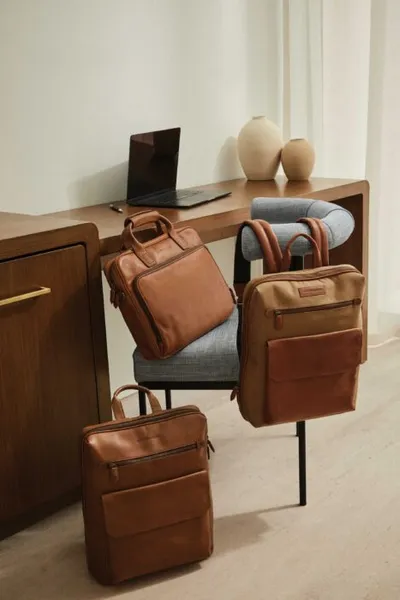 Three tan leather bags arranged on a chair and floor beside a wooden desk with a laptop