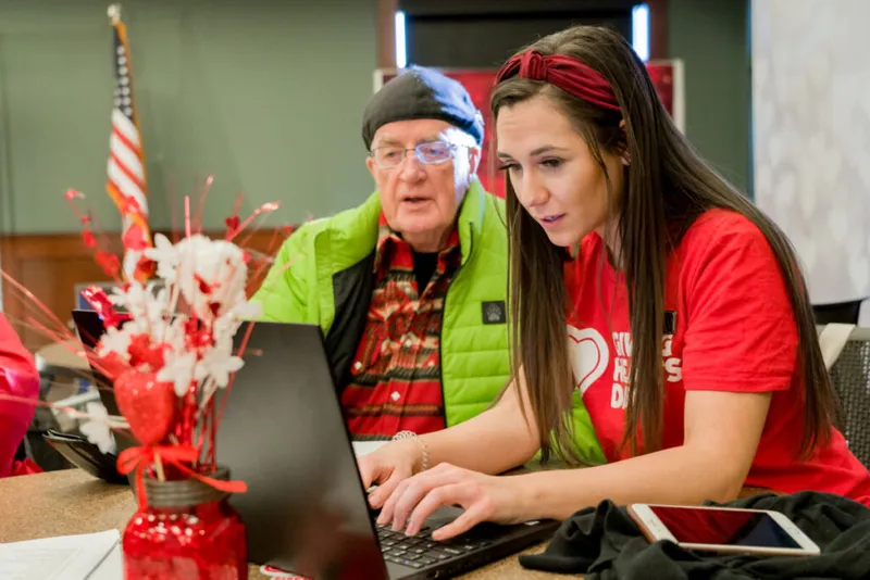 Young woman in red Giving Hearts Day shirt assists an older man at a laptop