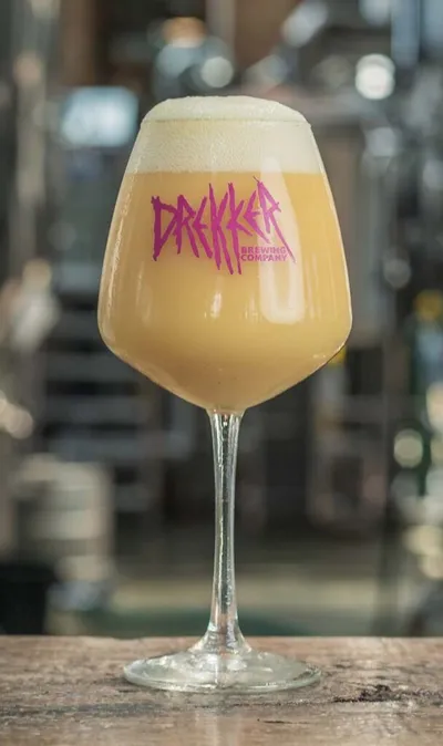 Hazy golden beer with thick white foam in a stemmed glass etched with Drekker Brewing Company logo