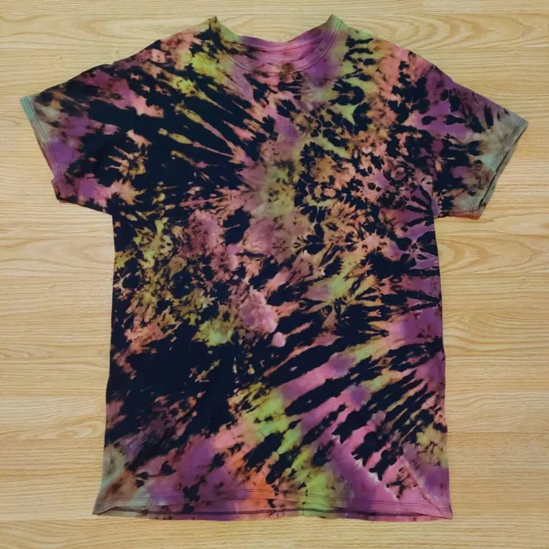 Flat-laid tie-dye T-shirt with black, pink, yellow-green, and purple swirling pattern on a wood surface