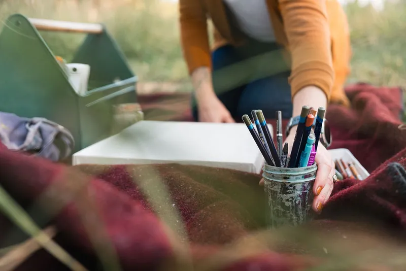 Artist reaching for a jar of pencils and pens beside a blank canvas on a plaid blanket outdoors
