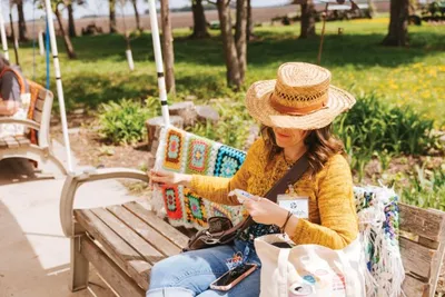Woman in straw hat and yellow cardigan examining a colorful crocheted piece on an outdoor bench