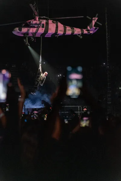 Performer descends a rope ladder from a pink helicopter above a dark concert crowd