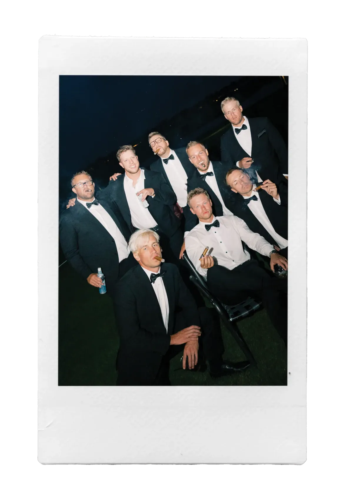 Group of eight men in black tuxedos and bow ties smoking cigars outdoors at night, captured on instant film