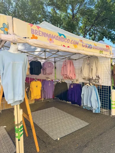 Outdoor market booth with colorful sweatshirts and tees hanging under a white canopy banner reading BrennaManoCo.com