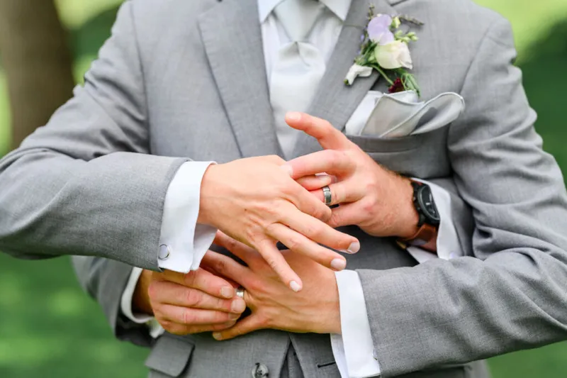Two grooms in gray suits exchanging wedding rings, one wearing a floral boutonniere