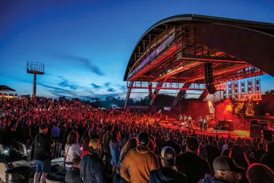 Large outdoor concert crowd facing a red-lit stage beneath a covered pavilion at dusk