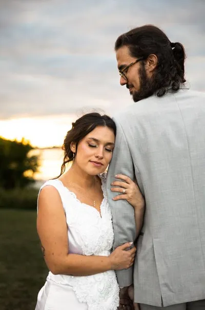 Bride in white lace gown resting her head on groom's gray suit jacket at sunset