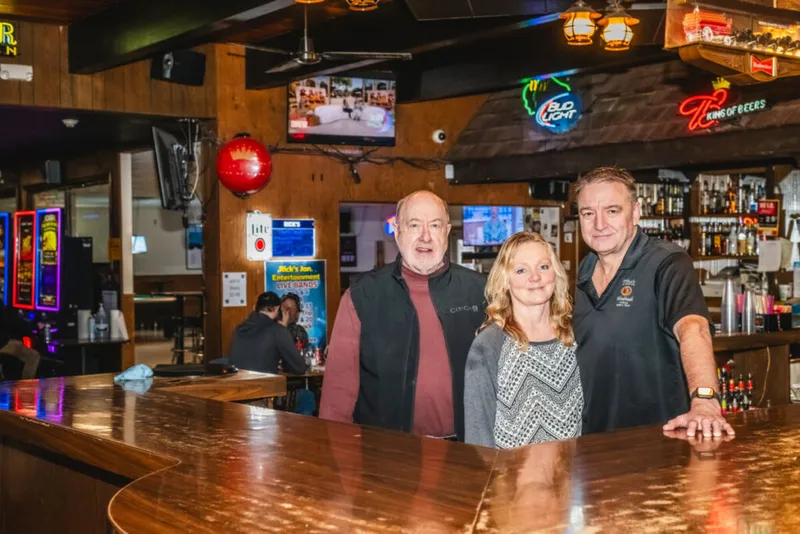 Two men and a woman smile behind a polished wood bar with neon beer signs and liquor shelves