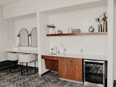 Stylish venue kitchenette with wood cabinets, floating shelf, and beverage fridge beside an arched mirror vanity area