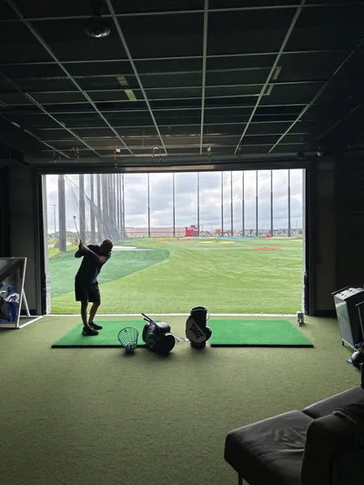 Man mid-swing at an indoor golf bay open to a driving range with golf bags nearby