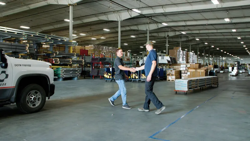 Two men shaking hands inside a large warehouse with stacked pallets and a white work truck nearby