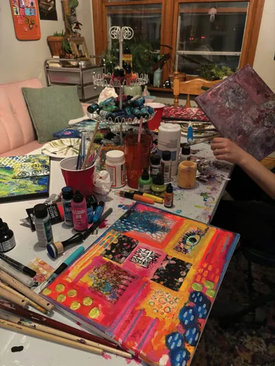 Cluttered art table covered in paint bottles, brushes, and colorful mixed-media canvases
