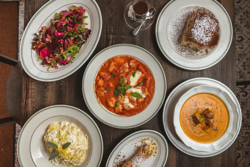 Overhead spread of six restaurant dishes including tomato stew, creamy risotto, beet salad, soup, and a dessert on a wood