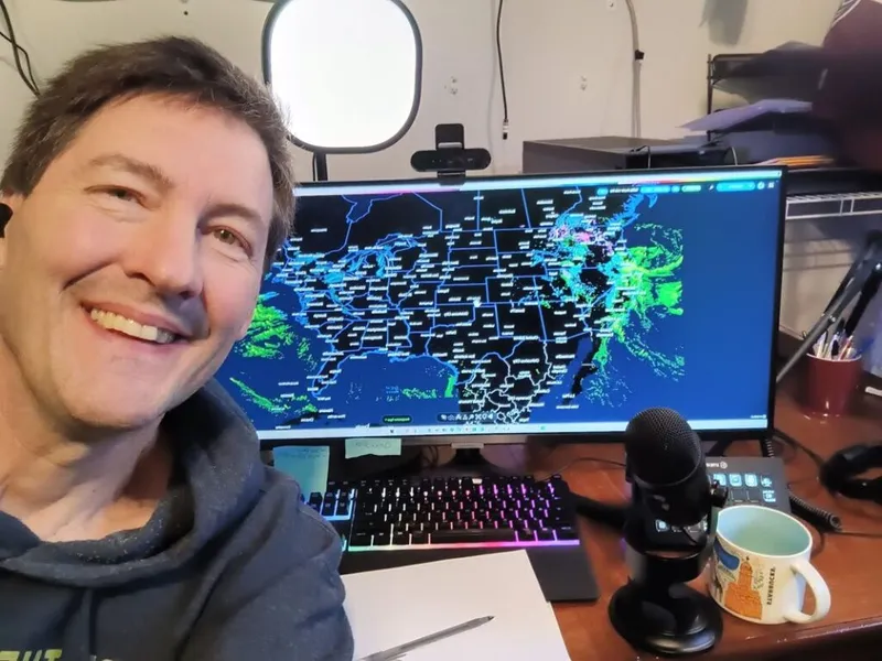 Smiling man at desk with radar map on wide monitor, microphone, and RGB keyboard