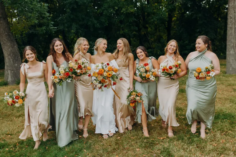 Bride in white gown laughing with seven bridesmaids in sage and champagne satin dresses holding orange and white bouquets
