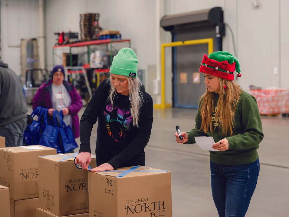 Two volunteers tape and check boxes labeled The Great North Pole in a warehouse, one wearing an elf hat