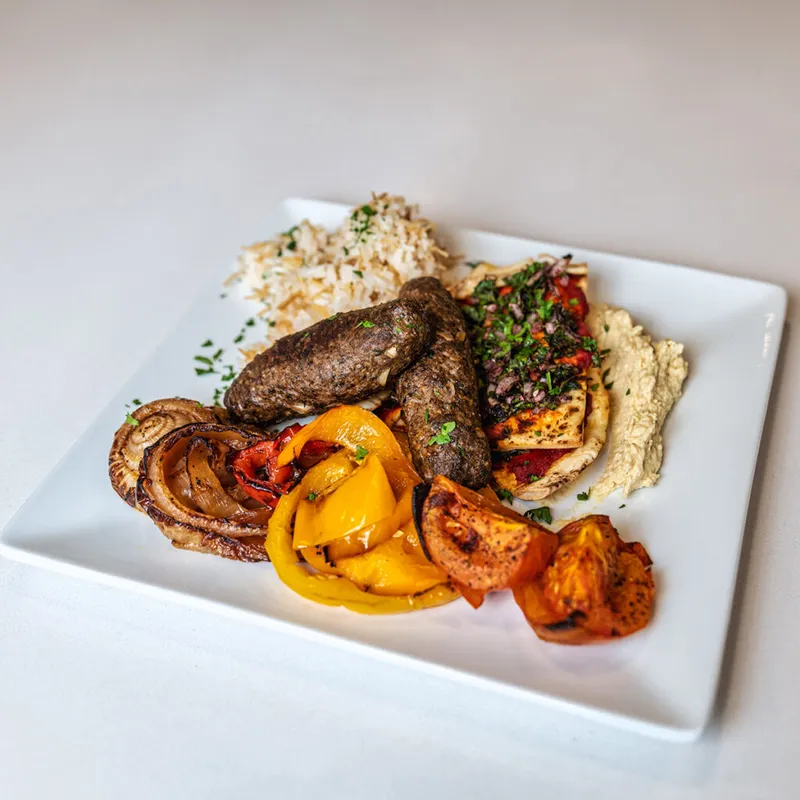 Grilled kofta kebabs with hummus, roasted peppers, mushrooms, and rice on a white square plate