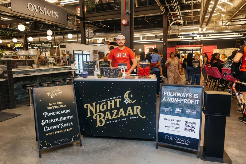 Man in red shirt stands behind a Folkways Night Bazaar booth inside a busy indoor market hall